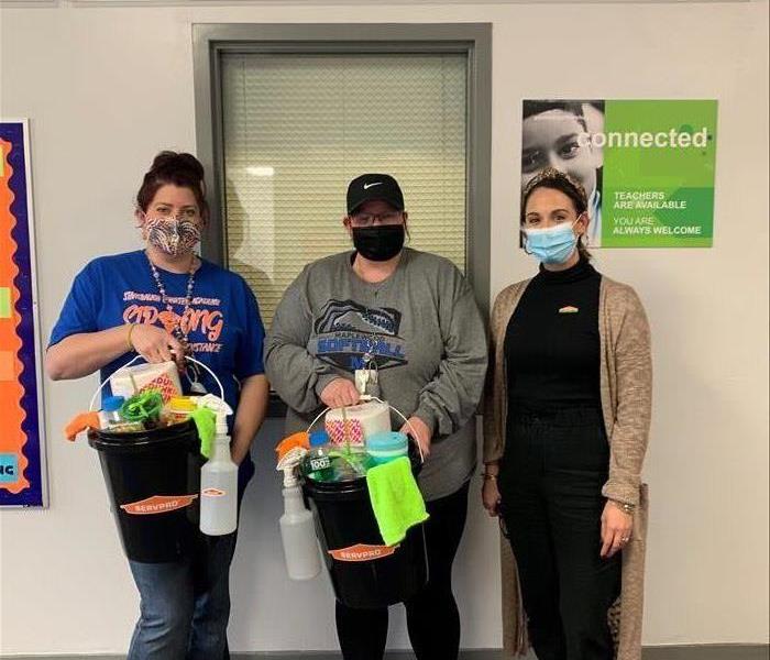 image of SERVPRO employee dropping off a bucket of cleaning supplies to two local teachers while smiling for a picture